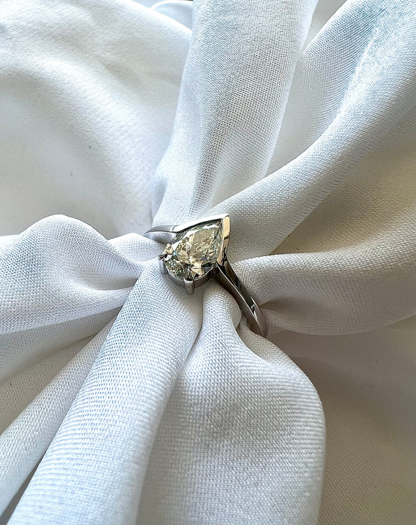 A close-up image of a diamond ring with a large central diamond set in a silver band, resting on a white fabric surface.