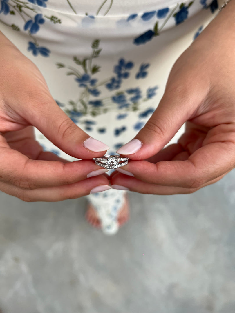 A close-up image of a person's hands holding a diamond engagement ring, with a blurred background that appears to be a floral patterned dress.