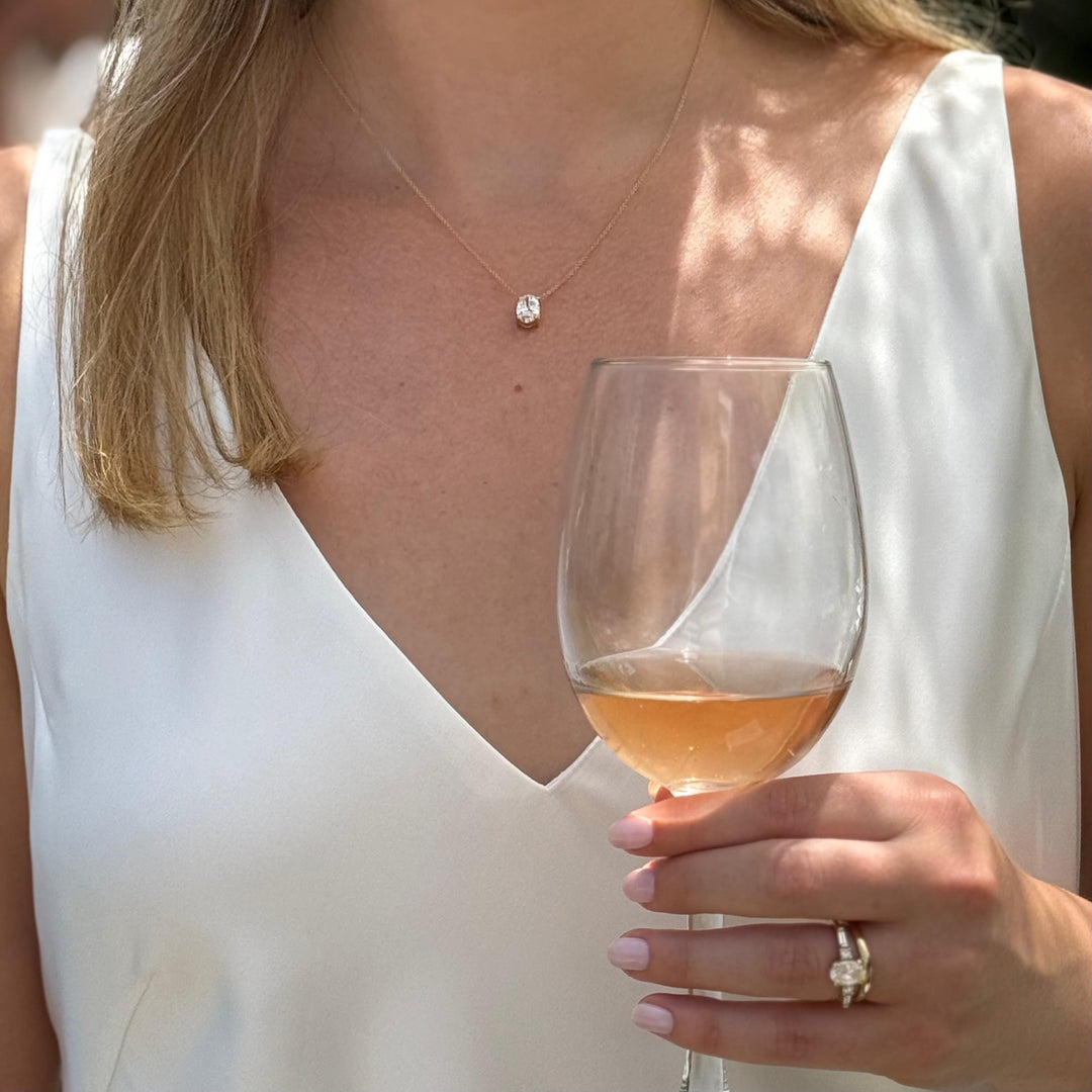 A woman in a white dress holds a glass of wine, with a necklace visible around her neck.