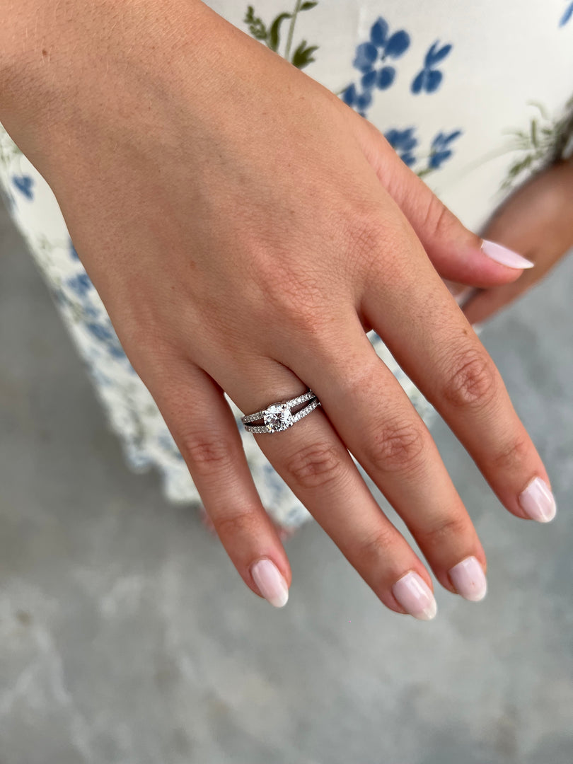 A close-up of a person's hand wearing a diamond engagement ring, with a floral patterned dress visible in the background.