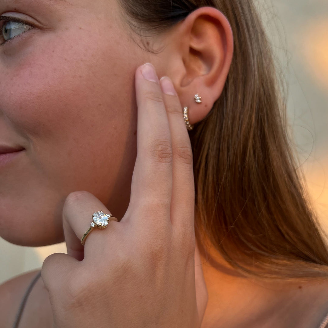 A close-up of a person's hand wearing a diamond engagement ring, with their earrings visible.