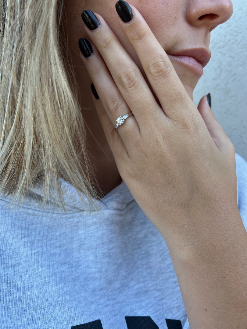 A close-up of a person's hand wearing a diamond engagement ring, with their fingers gently resting on their face.