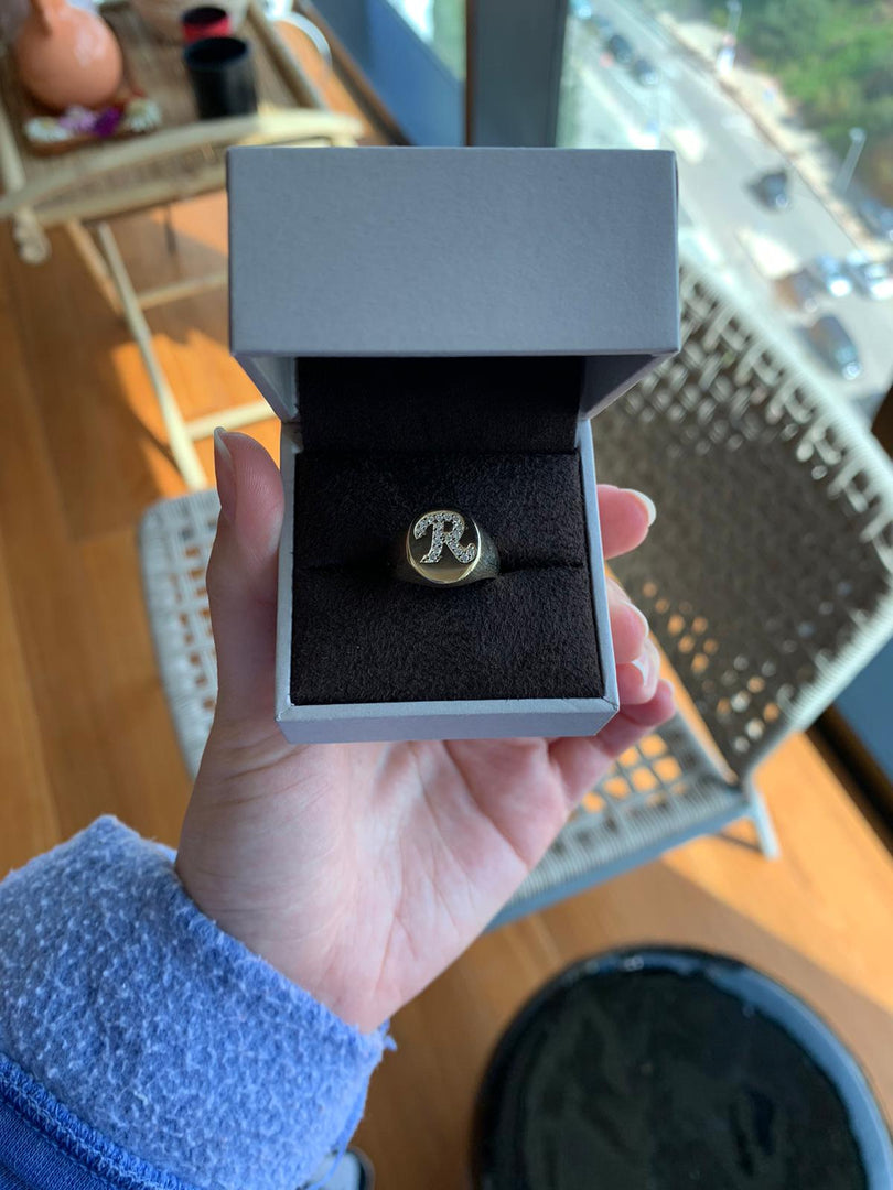 A person's hand holding a white jewelry box containing a gold ring with a large "R" on it.