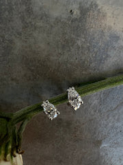 Two diamond earrings with a large central diamond and smaller diamonds on either side are displayed on a green stem against a textured gray background.