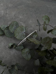 A silver bracelet with a diamond-encrusted design is resting on a bed of green leaves, creating a visually striking contrast between the metallic bracelet and the organic foliage.