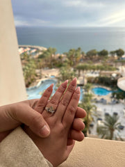 A close-up image of a person's hand holding a ring, with a scenic view of a beach and palm trees in the background.