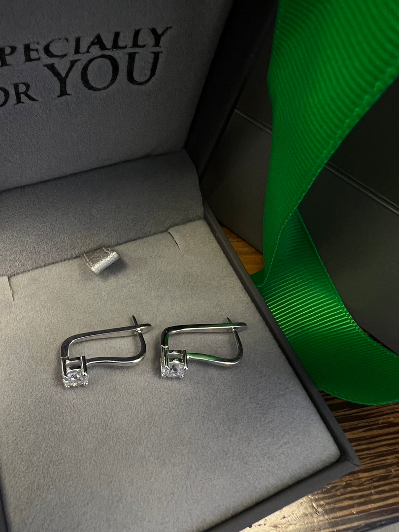 A pair of silver earrings with clear stones is displayed in a gray jewelry box with a green ribbon.